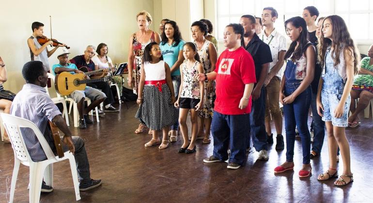 Cerca de doze alunos cantam de pé, em frente a orientador, que está sentado; ao lado, três pessoas com instrumentos musicais acompanham.