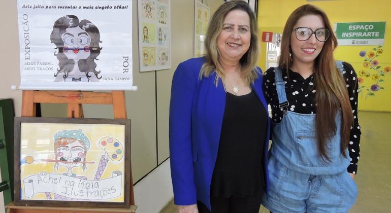 Duas mulheres ao lado de desenhos coloridos em exposição. 