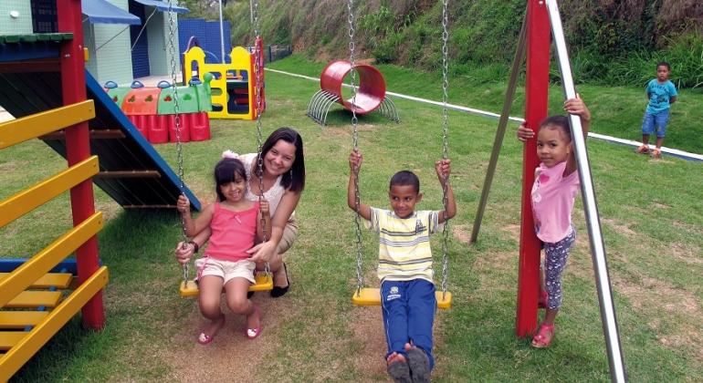 Três crianças brincam em balanço na área verde de Umei, auxiliadas por educadora infantil. 