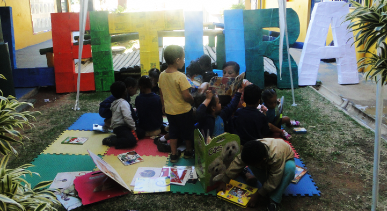 Com pedagogia ancorada na leitura, UMEI da região Leste de BH espalha letras em ‘tamanho gigante’ para encantar as crianças. Foto: Naara Dia Cerca de nove crianças manuseiam livros; atrás delas está escrito "Leitura" com letras coloridas e gigantes.