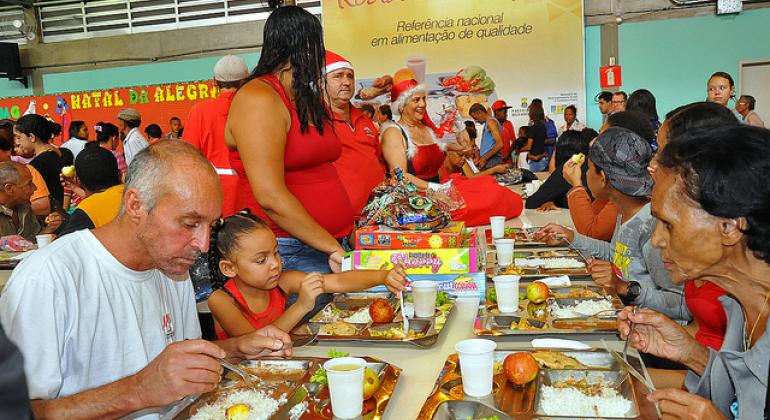 Cidadãos almoçam no restaurante popular enquanto pessoas vestidas de papai noel distribuem presentes