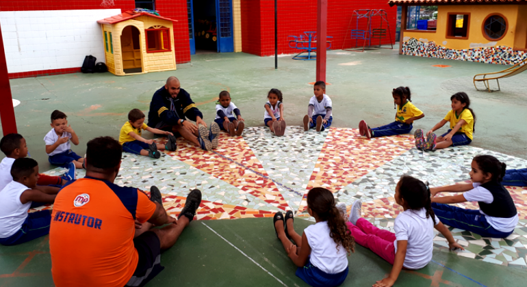 Prefeitura de Belo Horizonte oferece atividades físicas para crianças das Umeis e creches. Foto: Vilma Tomaz Treze crianças com idades entre 3 a 5 anos da Umei sentados, acompanhados por dois monitores, durante atividade física.