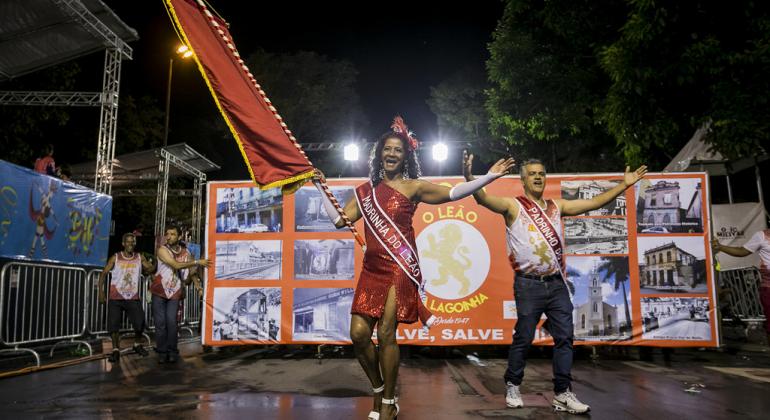 Passista de escola de samba é porta-bandeira, com vestido vermelho e faixa escrita "Madrinha do Leão". Ao lado, homem, representa o mestre-sala, e usa faixa com escrita "Padrinho do Leão". Os dois sambam na Avenida Afonso Pena à noite