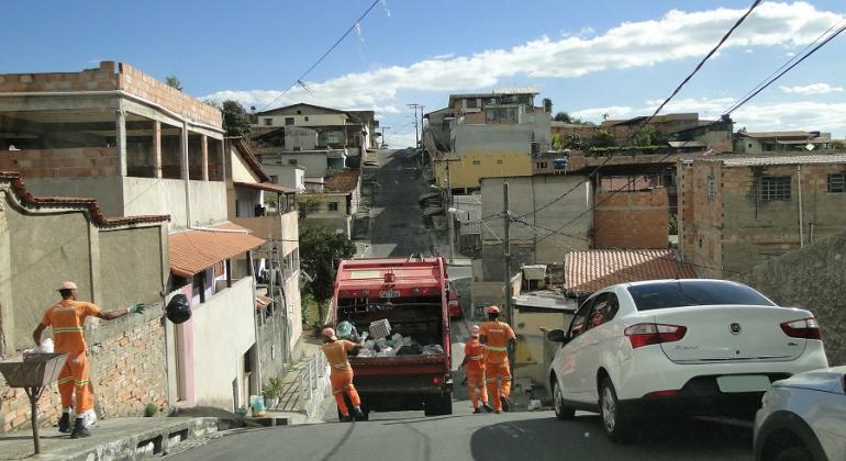 Caminhão de lixo com com quatro garis em rua íngreme durante o dia.