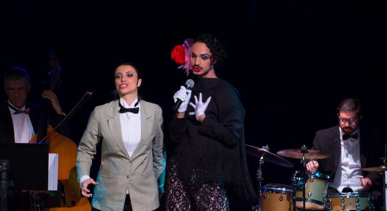 Cancioneiro Queer Dois atores interpretam uma canção. Ao fundo um baterista e um violoncelista