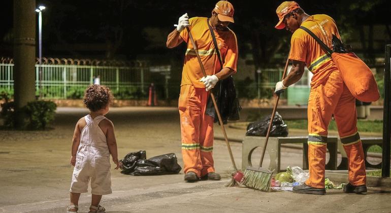 Dois garis varrem uma praça; ao lado uma criança caminha. 