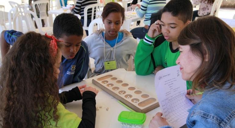 Crianças jogam jogo de tabuleiro, supervisionadas por educadora. Foto ilustrativa.