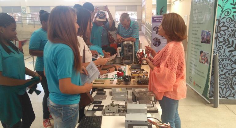 Mulher, de pé, em frente à mesa, atende a jovens em Semana da Orientação Profissional