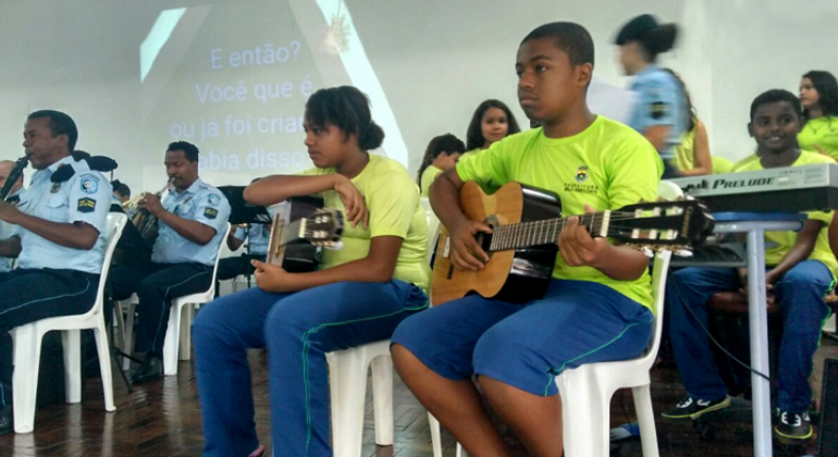 Alunos da Escola Municipal Dom Orione fizeram parte do Projeto Adote um Músico, através do qual aprendem música com a Guarda Municipal. Foto: Divulgação PBH Duas crianças alunas de escola municipal tocam violão; ao lado, guardas municipais tocam outros instrumentos.