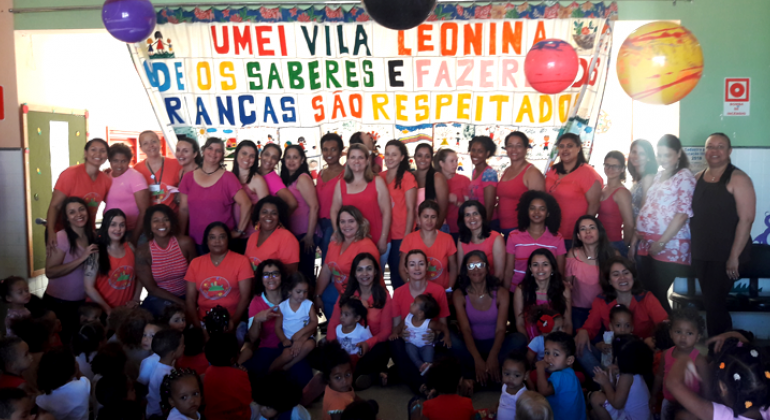 Equipe de cerca de 40 pessoas da Umei Vila Leonida se veste de rosa para conscientizar a todos sobre a importância da prevenção do câncer de mama. Ao fundo, uma grande faixa com os dizeres: UMEI Vila Leonina: onde os saberes e fazeres das crianças dão respeitados.