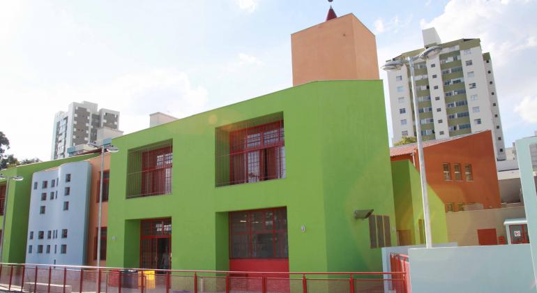 A foto mostra uma construção, paredes pintadas de verde claro e azul claro, grades de ferro pintadas de vermelho, duas janelas grandes e 2 portas de vidro. Atrás da construção vê-se dois prédios bem altos.