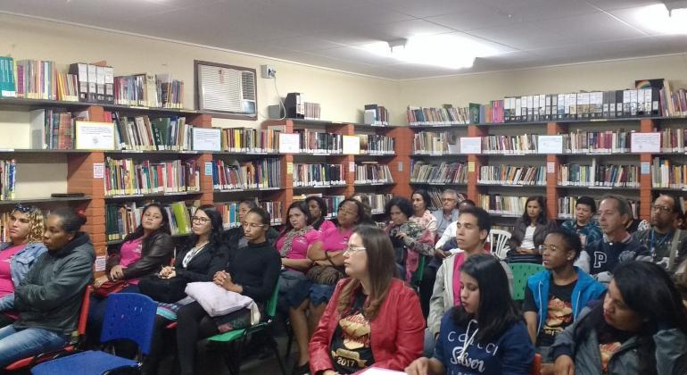 Grupo de cerca de vinte professores participa de aula em biblioteca. Estantes de livros ao fundo