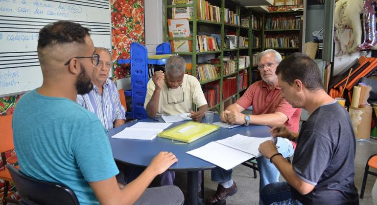 Centro de Convivência Carlos Prates é referência na inserção social de pessoas com sofrimento mental. Foto: Mia Nascimento/PBH Cinco pessoas sentadas em uma mesa com cadernos; ao fundo, vários livros em uma estante.