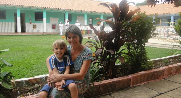 Alimentação saudável e cuidados com o meio ambiente são hábitos incentivados em creche conveniada da PBH. Foto: Naara Dias/PBH Mãe abraça filho sentada em canteiro com plantas, ao fundo, gramado e uma casa.