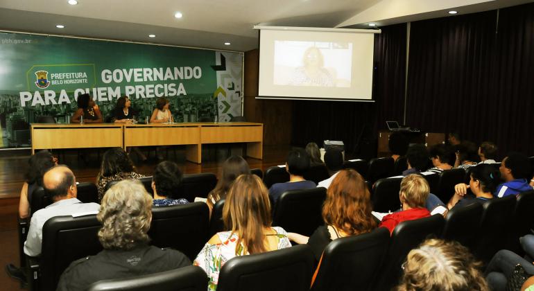 Seminário celebra os dez anos da Escola Integrada. Três pessoas estão sentadas em uma mesa, no palco. Na plateia há cerca de 30 pessoas, em duas fileiras de cadeiras.