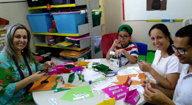 Novas salas de Atendimento Educacional Especializado fazem a diferença na política de Educação Inclusiva da PBH. Foto: Divulgação PBH Pais participam com o aluno de atividades propostas pela professora na sala de Atendimento Educacional Especializado; sentados à mesa, manipulam papéis coloridos e cola em bastão.