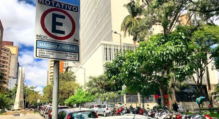PBH libera rotativos no Hipercentro aos sábados em dezembro Placa de estacionamento rotativo na rua Alvares Cabral, prédios e árvores ao fundo.
