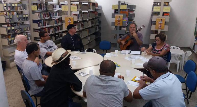 Nove pessoas, uma delas com violão, em torno de três mesas, fazendo parte do projeto "Feira da Poesia".