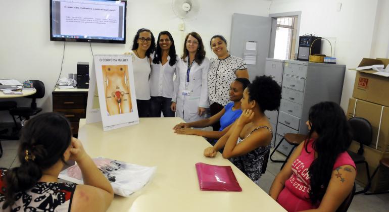 Mulheres do centro de saúde mostram desenho do corpo feminino, com destaque para os órgãos reprodutivos, para cidadãs sentadas: quatro mulheres estão de pé, e quatro estão sentadas. 