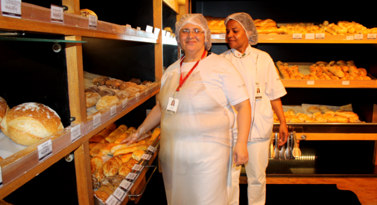 Duas mulheres com roupa branca, avental e touca, à frente de estante de padaria, com diversos pães.