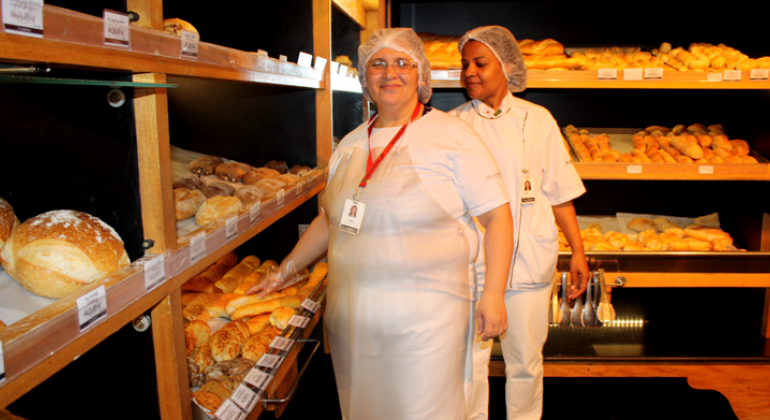 Iniciativa é fruto de parceria da Secretaria Municipal de Saúde, Ministério do Trabalho e Senac e beneficia 80 pessoas na capital mineira. Foto: Leila Porto Duas mulheres com roupa branca, avental e touca, à frente de estante de padaria, com diversos pães.