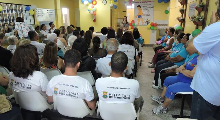 Mais de vinte profissionais do Centro de Saúde Padre Joaquim Maia, na Pampulha, reunidos na sala de espera do local, que comemora seu 10º aniversário; balões e adereços enfeitam o local. 