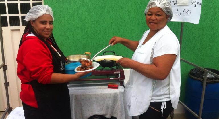 Duas cozinheiras servem tira-gostos em uma banca de feira.