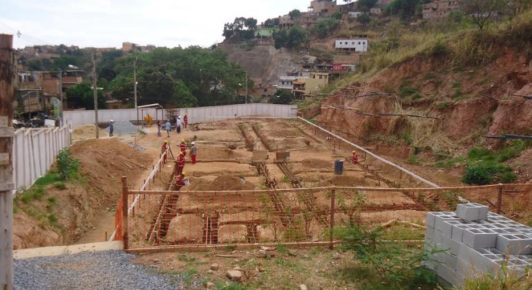 Terreno com marcação no chão para construção e tijolos de cimento; sete funcionários trabalham no local.