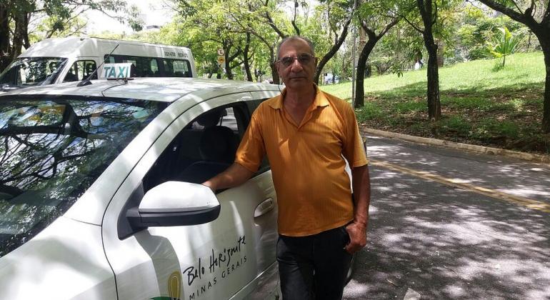 Taxista ao lado de táxi em rua arborizada, durante o dia.