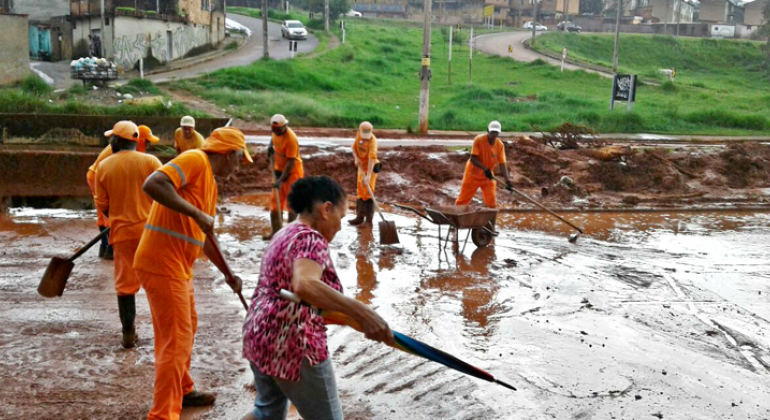Em toda a cidade, após os temporais, a Superintendência de Limpeza Urbana (SLU) já recolheu 330 toneladas de entulho. Foto: Divulgação/PBH Sete funcionarios da SLU realizam limpeza de rua após chuva e uma senhora passa pelo local.