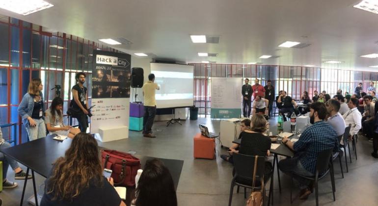 Foto: Divulgação PBH Homem dando palestra e mais de vinte pessoas assistindo em evento Hack a City