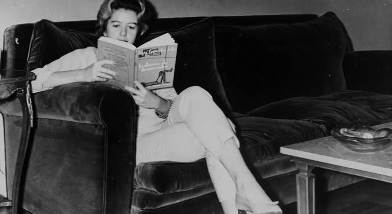 Sentada em um sofá, a poeta Hilda Hilst lê um livro. Foto preto e branco.