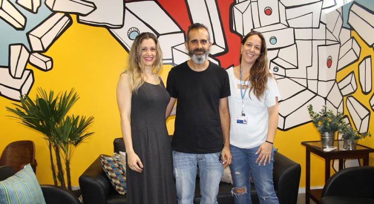 As paredes brancasde uma das salas da Secretaria Municipal de Planejamento ganharam vida e cor pelas mãos de um servidor e artista plástico. Foto: Aline Pereira/PBH Servidor e artista plástico Wagner Braccini acompanhado de duas servidoras, ao fundo, parede colorida pintada por ele.