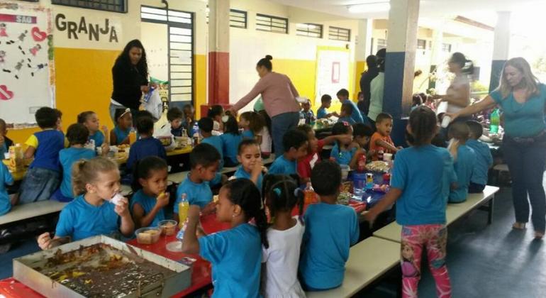 Foto: Arquivo PBH Mais de vinte crianças de seis anos lanchando, sentadas e supervisionadas por quatro profissionais de educação.