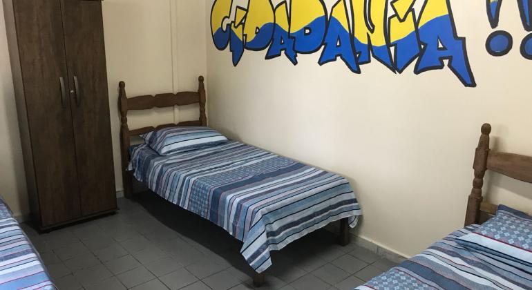 Quarto com três camas e um armário com grafite escrito "cidadania" em uma das paredes