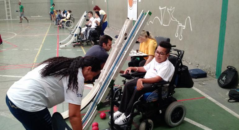 Criança com mobilidade reduzida em cadeira de rodas tem equipamento para jogar bocha colocado por técnica da Secretaria Municipal de Esportes.