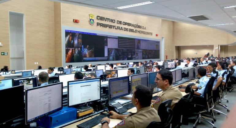 Sala com mais me 40 computadores, a maior parte deles ligados e com pessoas trabalhando. Na Parede, um grande telão com imagens. No alto do telão, os dizeres: Centro de Operações da Prefeitura de Belo Horizonte.