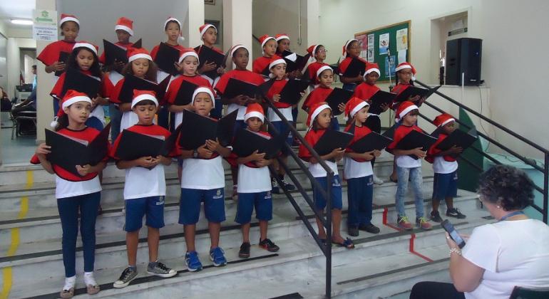 Mais de vinte crianças ensaiam em coral com gorro de Natal. 