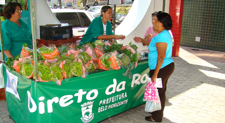 Dia 29 de dezembro, a PBH publicou edital de concorrência para a seleção de agricultores familiares que integrarão o Programa Direto da Roça. Foto: Divulgação PBH Duas mulheres observam alfaces em banca do Programa Direto da Roça.