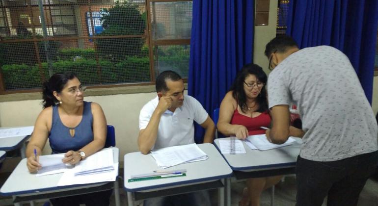 Três pessoas sentadas em mesas escolares; à frente, adulto preenche formulário, de pé, uma etapa da eleição para gestores escolares.