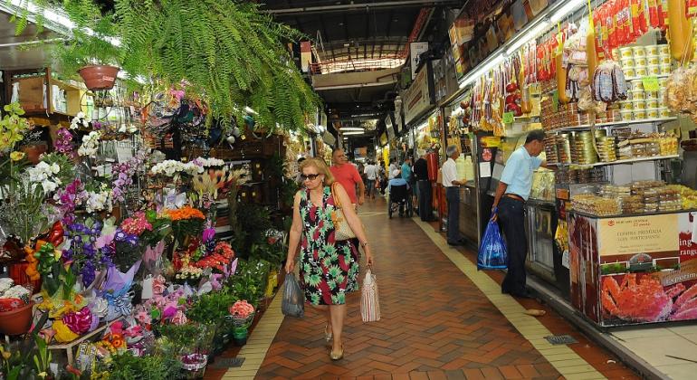 Mulher faz compras no Mercado Central, corredor tem flores de um lado e queijos de outro.