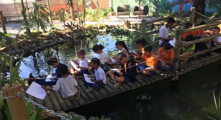 Mais de 12 crianças sentadas em uma ponte sob um laguinho.