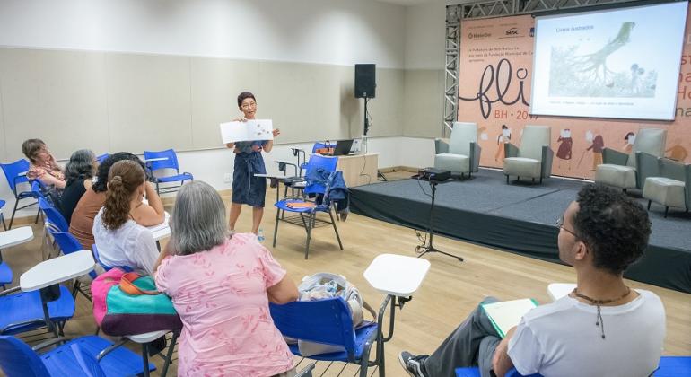 Foto: Divulgação PBH Mulher dá aula para quatro adultos, sentados em cadeiras.