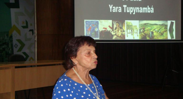 Palestrante Yara Tupinambá em pé e, logo atrás, uma imagem projetada de sua palestra.