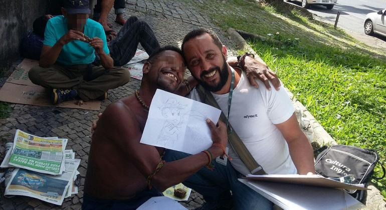 Morador em situação de rua abraça servidor e mostra desenho em folha branca. Os dois estão sentados no chão