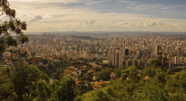 Vista panorâmica da cidade de Belo Horizonte, cm vegetação à frente, muitos prédios no meio e um céu com nuvens ao fundo.