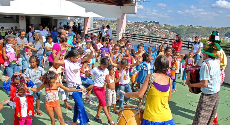 Cerca de 50 crianças participam de momento lúdico em escola durante as férias. Músicos se apresentam com tambores e fantasias.