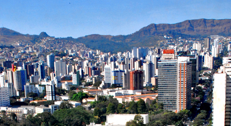 Cidade de Belo Horizonte, com muitos prédios e céu azul, durante o dia. Ao fundo, Serra do Curral.