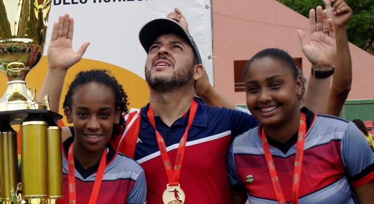 Duas garotas e um homem, todos com medalhas de ouro no peito, comemoram ao lado de troféu dourado.