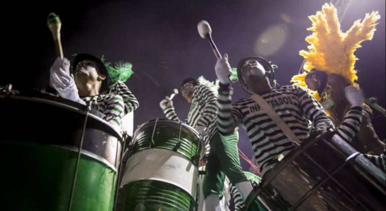 Componentes de Bateria de escola de samba vestidos de ladrão batucam em tambores em formato de lata de lixo